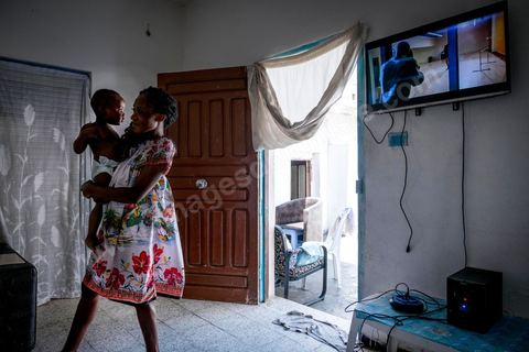 24 juin 2017 : Océane et son fils dans le salon. Chez Océane, migrante Ivoirienne à Tunis dans un quartier populaire. Elle vit dans une maison avec 2 chambres qu'elle partage avec son fils Eden et cinq autres ivoiriennes. Océane est arrivée en Tunisie en 2010 comme étudiante et est depuis 5 ans en situation irrégulière, ne lui permettant plus de travailler légalement. Il y a deux ans Eden est né, ce jeune garçon plein de joie est devenu sa raison de vivre, elle souhaite trouver un endroit pour vivre normalement et élever son fils. “J’estime que pour vivre bien Tunisie, il me faudrait entre 800 et 1000 dinars. Généralement, je suis plutôt à 350. Entre les couches et la nourriture, les dépenses sont rapides.” La jeune femme travaille de façon irrégulière : elle a quelques patrons qui l’appellent, selon leur besoin, pour faire le ménage chez eux. Une journée lui rapporte entre 25 et 30 dinars.  