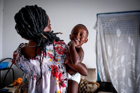 24 juin 2017 : Océane et son fils dans le salon. Chez Océane, migrante Ivoirienne à Tunis dans un quartier populaire. Elle vit dans une maison avec 2 chambres qu'elle partage avec son fils Eden et cinq autres ivoiriennes. Océane est arrivée en Tunisie en 2010 comme étudiante et est depuis 5 ans en situation irrégulière, ne lui permettant plus de travailler légalement. Il y a deux ans Eden est né, ce jeune garçon plein de joie est devenu sa raison de vivre, elle souhaite trouver un endroit pour vivre normalement et élever son fils. “J’estime que pour vivre bien Tunisie, il me faudrait entre 800 et 1000 dinars. Généralement, je suis plutôt à 350. Entre les couches et la nourriture, les dépenses sont rapides.” La jeune femme travaille de façon irrégulière : elle a quelques patrons qui l’appellent, selon leur besoin, pour faire le ménage chez eux. Une journée lui rapporte entre 25 et 30 dinars.  