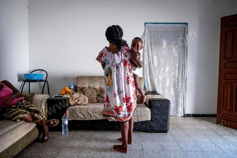 24 juin 2017 : Océane et son fils dans le salon. Chez Océane, migrante Ivoirienne à Tunis dans un quartier populaire. Elle vit dans une maison avec 2 chambres qu'elle partage avec son fils Eden et cinq autres ivoiriennes. Océane est arrivée en Tunisie en 2010 comme étudiante et est depuis 5 ans en situation irrégulière, ne lui permettant plus de travailler légalement. Il y a deux ans Eden est né, ce jeune garçon plein de joie est devenu sa raison de vivre, elle souhaite trouver un endroit pour vivre normalement et élever son fils. “J’estime que pour vivre bien Tunisie, il me faudrait entre 800 et 1000 dinars. Généralement, je suis plutôt à 350. Entre les couches et la nourriture, les dépenses sont rapides.” La jeune femme travaille de façon irrégulière : elle a quelques patrons qui l’appellent, selon leur besoin, pour faire le ménage chez eux. Une journée lui rapporte entre 25 et 30 dinars.  