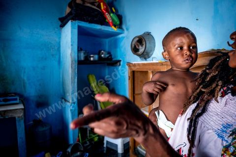 24 juin 2017 : Océane et son fils dans la cuisine. Chez Océane, migrante Ivoirienne à Tunis dans un quartier populaire. Elle vit dans une maison avec 2 chambres qu'elle partage avec son fils Eden et cinq autres ivoiriennes. Océane est arrivée en Tunisie en 2010 comme étudiante et est depuis 5 ans en situation irrégulière, ne lui permettant plus de travailler légalement. Il y a deux ans Eden est né, ce jeune garçon plein de joie est devenu sa raison de vivre, elle souhaite trouver un endroit pour vivre normalement et élever son fils. “J’estime que pour vivre bien Tunisie, il me faudrait entre 800 et 1000 dinars. Généralement, je suis plutôt à 350. Entre les couches et la nourriture, les dépenses sont rapides.” La jeune femme travaille de façon irrégulière : elle a quelques patrons qui l’appellent, selon leur besoin, pour faire le ménage chez eux. Une journée lui rapporte entre 25 et 30 dinars.  