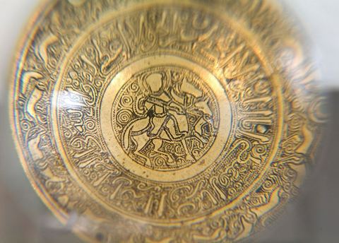 PLATEAU DE BALANCE AU CAVALIERMésopotomie (Irak),12  - 13  siècle. Cuivre et traces d'argent.La pièce, percée de trois trous de suspension, est finement ciselée. Au centre, un cavalier vu de face, à la figure nimbée d'une auréole, accompagné de son lévrier. Il est entouré de spires renforçant la position dynamique du cheval et du lévrier. Tout autour, une dernière frise en caractère cursif 'naskhi', aux courbures accentuées, tapissée des mêmes spires. Elle livre, partiellement, un verset du Coran se référant au thème de la 'juste balance'. Sur la dernière frise, une sphinge, corps de félin ailé et tête de femme, et une suite d'animaux passant, liés au thème de la chasse: lion, guépard, cerfs, antilope, lévrier.Si la relation est évidente entre la balance et le passage du Coran, l'association entre divers thèmes, tels que la mesure, la parole divine, une créature fabuleuse, un cavalier auréolé et une scène de chasse paraît incongrue. Elle trouve cependant sa cohérence dans la dimension ornementale d'une oeuvre plaisante au regard, et la réussite d'un beau mouvement dynamique. Dans de nombreuses pièces de l'art arabe et islamique, il ne faut chercher ni symbole, ni 'horreur du vide', mais souvent, simplement, un décor pour le décor, au sens le plus fort d'un art pour l'art