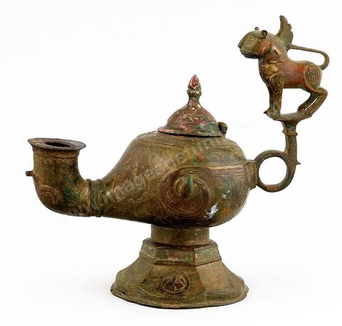 LAMPE À HUILE AU LION AILÉ.Khorassan, 12  siècle.La représentation du merveilleux puise sa source dans les croyances de l'Arabie pré-islamique, les mythes de l'Antiquité Classique et iranienne auxquels se sont greffés les apports de l'Asie Centrale et la fascination qu'exerçaient les récits de voyage. Phénix, harpie, griffon, pégase, dragon, waq-waq (arbre à figures humaines) ornaient objets, textiles et manuscrits. Le sphinx, un lion à figure humaine, avec ou sans ailes, y prenait une bonne place. Les lampes à huile en métal sont, le plus souvent, munies d'une figure d'oiseau rehaussant le poucier.Sur cette vigoureuse lampe à huile, les ailes, symbole de lumière et de survie, le lion, symbole de majesté et de maîtrise, s'associent, en ronde-bosse, pour conférer à la lampe une sorte de valeur 'magique', évoquant force et éternité. - Musée Lella Hadria à Djerba Explore