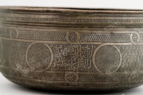 BASSIN EN CUIVRE ÉTAMÉ. Egypte/Syrie, 15 - 16  siècle.À l'heure de la chute de la dynastie mamelouke (1258-1512) et de l'annexion de la Syrie et de l'Egypte à l'Empire ottoman, le manque de métal précieux incita les artisans à enrichir les surfaces des pièces de décors ciselés très denses. Ils devaient couvrir, comme sur ce bassin, jusqu'aux parties les moins visibles, tel que le fond extérieur. L'étamage qui couvre la couleur cuivrée et l'incrustation d'une pâte bitumineuse noire, devaient apporter les effets d'éclat et de contraste, obtenus habituellement par l'argent. - Musée Lella Hadria à Djerba Explore