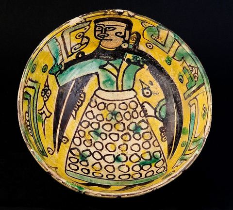 COUPE DE LA DAME EN CAPE NOIREKhorassan, Nichapour, 10 - 11  siècleUn personnage féminin en vêtement ocellé et cape noire à fausses manches pointues, tient à  la main un objet difficile à identifier (hache, miroir ou parchemin).Le thème de la dame debout, à l'oeil étiré (mais vu de profil), tenant un objet, se rencontre dans la céramique 'tunisienne' du 10 siècle. C'est le décor central ornant deux plats dits Dame de Sabra, découverts sur le site fatimide de Sabra Mansouriya et conservés au Musée des Arts Islamiques à Raqqada (Kairouan).