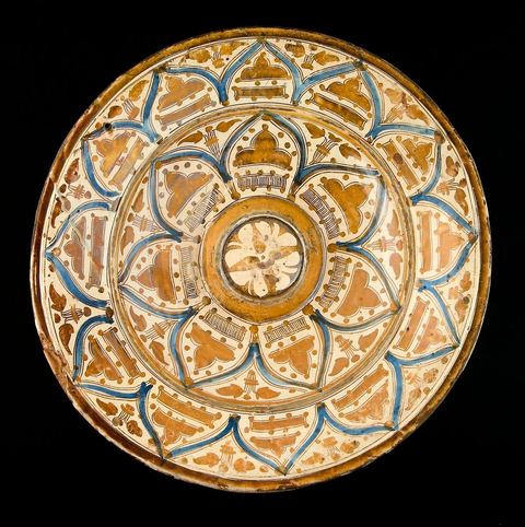 GRAND PLAT RAYONNANT BLEU ET OR. Espagne, Valence, (art hispano-mauresque), 16  siècle.Héritière des traditions andalouses, la céramique en lustre d'or, la loza dorada, s'est maintenue en Espagne après la Reconquista chrétienne. Monumentale et somptueuse, marquée d'un nouvel essor, elle fut recherchée tant en Europe qu'en Tunisie ou en Egypte. Son mode de cuisson, en trois étapes, est le même qu'en Orient. Sa composition chimique reprend, globalement, celle donnée par Abou Al Kacim de Kachan (13  siècle). Un ton plus doré que cuivré témoigne, en général, d'une plus importante proportion de l'or.  Sur ce plat, autour d'un motif floral central, traité en réserve, évoluent deux registres concentriques reproduisant le même décor. Une série d'arcs blancs, en ogives, cernés d'un double filet or, séparés par des bouquets, sont meublés de plus petits 'arcs trilobés' pleins et de bandes hachurées ou pleines, ornées de perles. L'ensemble évoque une large composition florale en couronne. Le registre le plus large est divisé en douze arcatures et le plus petit en sept, alors qu'une juste proportion géométrique n'en aurait donné que six. Outre la valeur religieuse et astrale du '12' et du '7', aussi bien en chrétienté qu'en Islam, cette division, accentuée par la couleur bleue, renforce l'effet de dynamique et de chatoiement déjà obtenus par les reflets du lustre. L'association du bleu et de l'or est caractéristique des plus belles séries de la céramique hispano-mauresque. On la retrouve dans les oeuvres  les plus somptueuses de l'art andalous et ifrikiyen: le cadre extérieur du 'Mihrab' de la Grande Mosquée de Cordoue en Espagne et le célèbre manuscrit en parchemin du 'Coran bleu' de Kairouan en Tunisie - Lalla Hadria Museum - Djerba Explore - Djerba