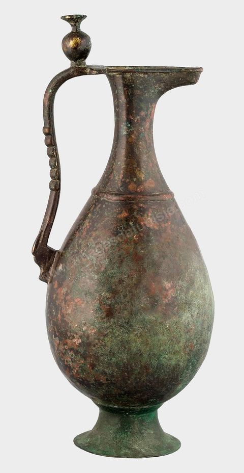 AIGUIÈRE EN BRONZEAttribuée à la Syrie, période omeyyade, 8  siècle.Les bronziers et dinandiers des premières périodes de l'art arabe se contentaient le plus souvent de proposer des aiguières de modèles piriformes, vigoureux, bien proportionnés et sans ornements. La partie supérieure du col, le bec, une reprise d'une forme sassanide, plus ancienne, évoque une tête d'animal. L'anse incurvée, directement attachée à la lèvre, rehaussée d'un poucier en forme de grenade – signe d'abondance et de prospérité -, est ornée de six perles en son milieu. L'usage de ce genre d'aiguière est souvent associé au service du vin.