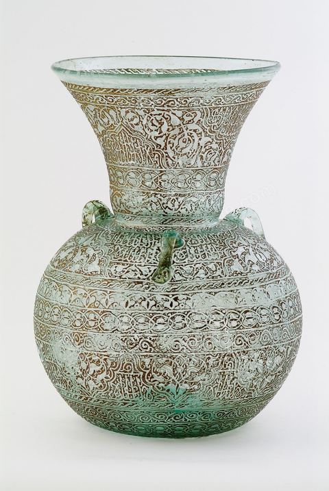 LAMPE DE MOSQUÉE EN VERRE GRAVÉ ET DORÉ.Syrie ottomane, 19  siècle.Cette lampe de mosquée à trois anses, à panse globulaire et haut col évasé reproduit un modèle classique  connu en Egypte ou en Syrie durant la période mamelouke (14 siècle). La même forme caractérise une poterie du 10 siècle, trouvée à Raqqada en Ifrikya (Tunisie). Elle inspirera les céramistes d'Iznik en Turquie (16 siècle) et plus tard les verriers d'Europe et de Syrie (19 siècle).À haute valeur symbolique, on la retrouve reproduite dans les portières de mosquée, les tapis de prière de Perse ou d'Anatolie où elle suggère un renvoi à un célèbre passage du Coran. Chapitre de La Lumière V.35. Voir tapis adjacent.