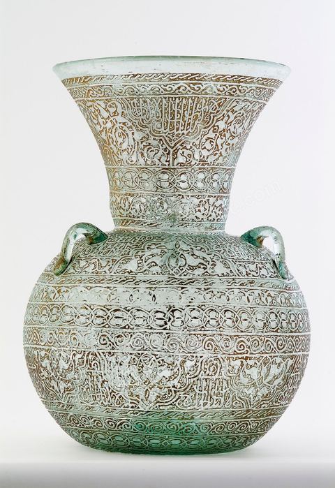 LAMPE DE MOSQUÉE EN VERRE GRAVÉ ET DORÉ.Syrie ottomane, 19  siècle.Cette lampe de mosquée à trois anses, à panse globulaire et haut col évasé reproduit un modèle classique  connu en Egypte ou en Syrie durant la période mamelouke (14 siècle). La même forme caractérise une poterie du 10 siècle, trouvée à Raqqada en Ifrikya (Tunisie). Elle inspirera les céramistes d'Iznik en Turquie (16 siècle) et plus tard les verriers d'Europe et de Syrie (19 siècle).À haute valeur symbolique, on la retrouve reproduite dans les portières de mosquée, les tapis de prière de Perse ou d'Anatolie où elle suggère un renvoi à un célèbre passage du Coran. Chapitre de La Lumière V.35. Voir tapis adjacent.