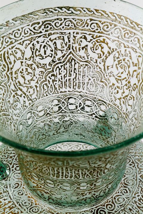 LAMPE DE MOSQUÉE EN VERRE GRAVÉ ET DORÉ.Syrie ottomane, 19  siècle.Cette lampe de mosquée à trois anses, à panse globulaire et haut col évasé reproduit un modèle classique  connu en Egypte ou en Syrie durant la période mamelouke (14 siècle). La même forme caractérise une poterie du 10 siècle, trouvée à Raqqada en Ifrikya (Tunisie). Elle inspirera les céramistes d'Iznik en Turquie (16 siècle) et plus tard les verriers d'Europe et de Syrie (19 siècle).À haute valeur symbolique, on la retrouve reproduite dans les portières de mosquée, les tapis de prière de Perse ou d'Anatolie où elle suggère un renvoi à un célèbre passage du Coran. Chapitre de La Lumière V.35. Voir tapis adjacent.
