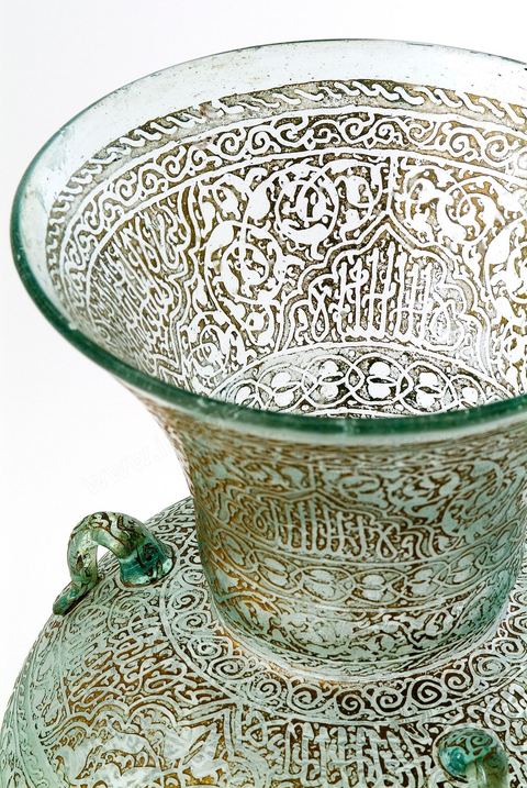 LAMPE DE MOSQUÉE EN VERRE GRAVÉ ET DORÉ.Syrie ottomane, 19  siècle.Cette lampe de mosquée à trois anses, à panse globulaire et haut col évasé reproduit un modèle classique  connu en Egypte ou en Syrie durant la période mamelouke (14 siècle). La même forme caractérise une poterie du 10 siècle, trouvée à Raqqada en Ifrikya (Tunisie). Elle inspirera les céramistes d'Iznik en Turquie (16 siècle) et plus tard les verriers d'Europe et de Syrie (19 siècle).À haute valeur symbolique, on la retrouve reproduite dans les portières de mosquée, les tapis de prière de Perse ou d'Anatolie où elle suggère un renvoi à un célèbre passage du Coran. Chapitre de La Lumière V.35. Voir tapis adjacent.