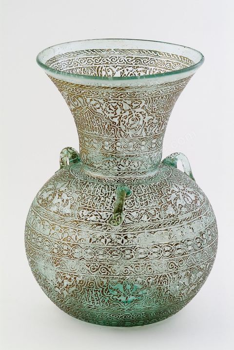 LAMPE DE MOSQUÉE EN VERRE GRAVÉ ET DORÉ.Syrie ottomane, 19  siècle.Cette lampe de mosquée à trois anses, à panse globulaire et haut col évasé reproduit un modèle classique  connu en Egypte ou en Syrie durant la période mamelouke (14 siècle). La même forme caractérise une poterie du 10 siècle, trouvée à Raqqada en Ifrikya (Tunisie). Elle inspirera les céramistes d'Iznik en Turquie (16 siècle) et plus tard les verriers d'Europe et de Syrie (19 siècle).À haute valeur symbolique, on la retrouve reproduite dans les portières de mosquée, les tapis de prière de Perse ou d'Anatolie où elle suggère un renvoi à un célèbre passage du Coran. Chapitre de La Lumière V.35. Voir tapis adjacent.