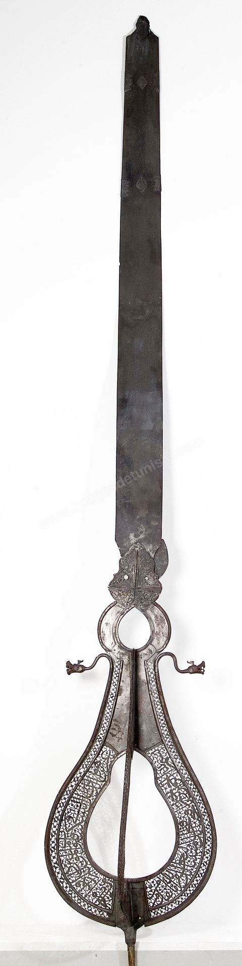 ÉTENDARD DE CÉRÉMONIE RELIGIEUSE. Perse, safavide,  daté de 1658 - 1659, mis en main-morte en 1696-7Acier décoré en ajour où s'inscrit en plein un extrait de la Sourate 48, verset 4, du Coran:  'Lui qui fait descendre la sérénité au coeur des croyants pour que, déjà croyants, ils grandissent dans la croyance.' 'À Dieu armées des cieux et de la terre et Dieu est connaissant et Sage '.L'exégèse relie ce passage du Coran tiré du Chapitre Al Fath, 'le Succès' ou 'la Conquête', à une bataille menée par le prophète Mohammad contre les païens au sujet, semble-t-il, de la prise de la Mecque. Prêts à se déployer, gueules ouvertes et dents acérées, deux dragons veillent à la protection de l'étendard. D'origine chinoise le thème du dragon, connu dans les cultures turques, était très en vogue en Perse durant la période safavide (16  - 18  siècle). La Porte dite du 'Talisman' à Bagdad, disparue aujourd'hui, était déjà ornée d'un impressionnant couple de dragons, symbole de majesté et de protection. (13 siècle)