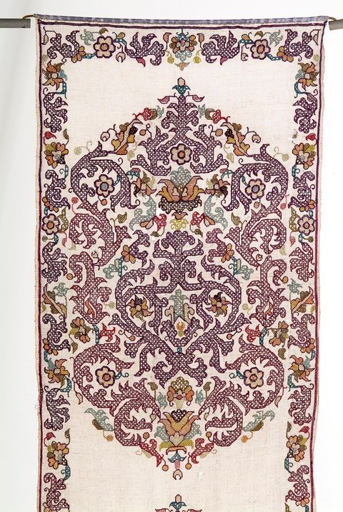 TENTURE À DÉCOR DE RINCEAUX FLEURIS. Alger, 18  siècle.Lin brodé de fil de soie polychrome à dominante violette. Aux extrémités, une rivière travaillée à  l'aiguille et exécutée en soie grège. En vogue à Alger, la teinte violette, issue du bois de campêche (importé d'Amérique), était très rare dans la broderie méditerranéenne. Chef-d'oeuvre de la broderie algérienne, cet ouvrage emprunte, en partie, son répertoire floral à l'herbier de la Turquie ottomane et sa composition, en gros rinceaux fleuris, à la Renaissance italienne. Une trace lointaine des puissantes compositions de l'art maghrébin almohade (12 - 13  siècle) n'est pas exclue.Recherchée en Europe, l'exemplaire le plus ancien de la broderie d'Alger se trouve en France. Il pare aujourd'hui 'La vierge noire' de Notre-Dame-du-Pilier à la Cathédrale de Chartres.