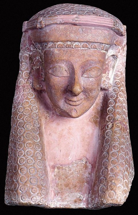 Masque de femme ( protomé ) en terre cuite. Le visage est ovale, les yeux en amande, la bouche souriante et le menton est agrémenté d’une fossette. Un klaft maintenu sur le front par un diadème recouvre sa tête. Placés dans les tombes, ces masques sont censés protéger l’âme des défunts. Les plus anciens ont un style égyptisant très marqué et ne sont pas sans évoquer les momies (H.‑31‑cm, nécropole de Dermech à‑Carthage, fin du VIe siècle av.J.-C., musée du Bardo ). Section punique - musée du Bardo